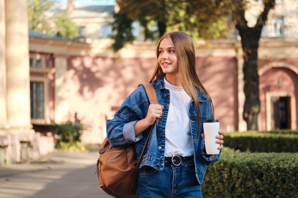 Pretty smiling student girl in denim jacket with backpack and coffee on study break for GRE tutoring at American Admissions